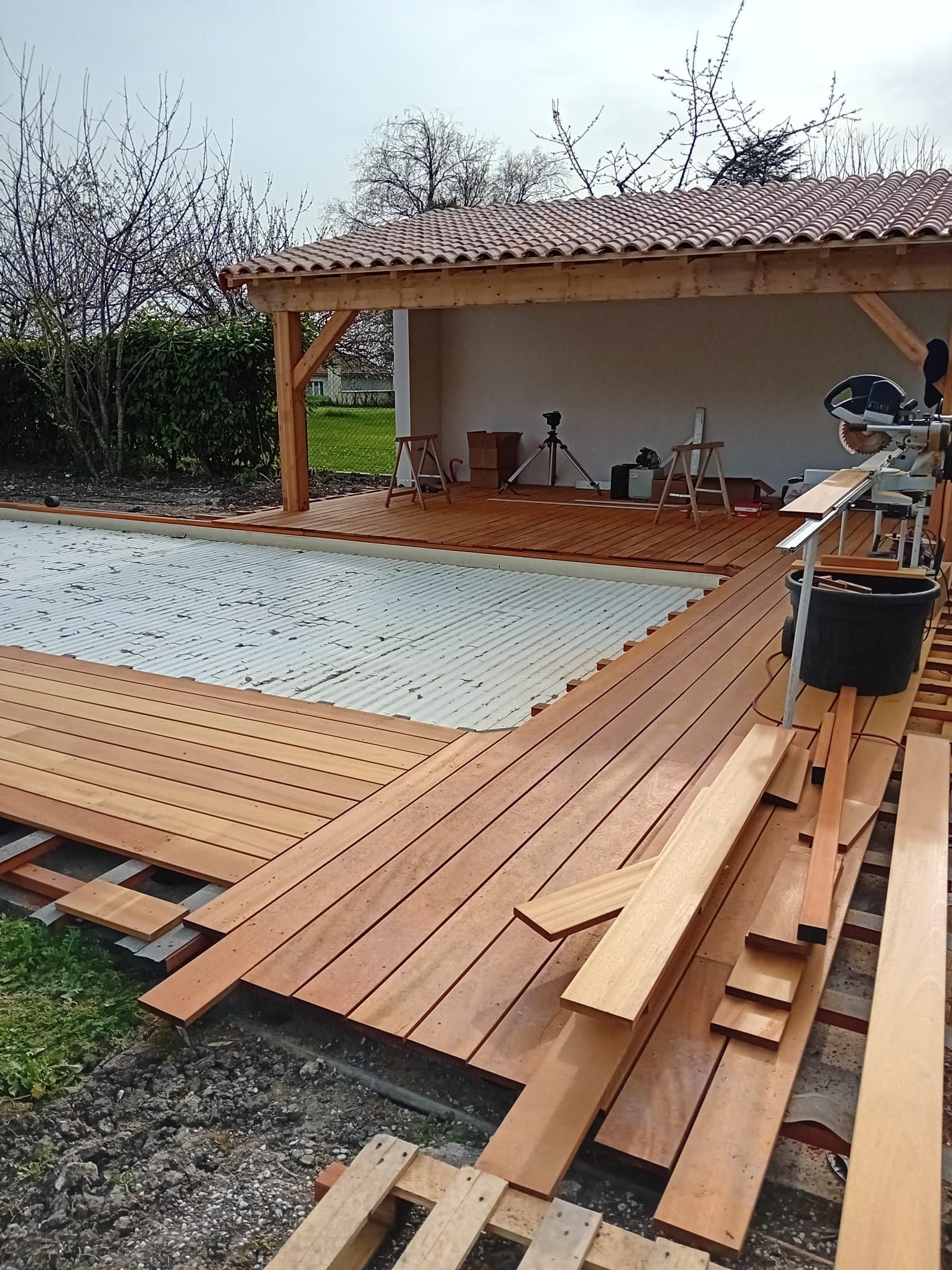 Une terrasse en bois est en construction près d'une piscine, sous un patio couvert.