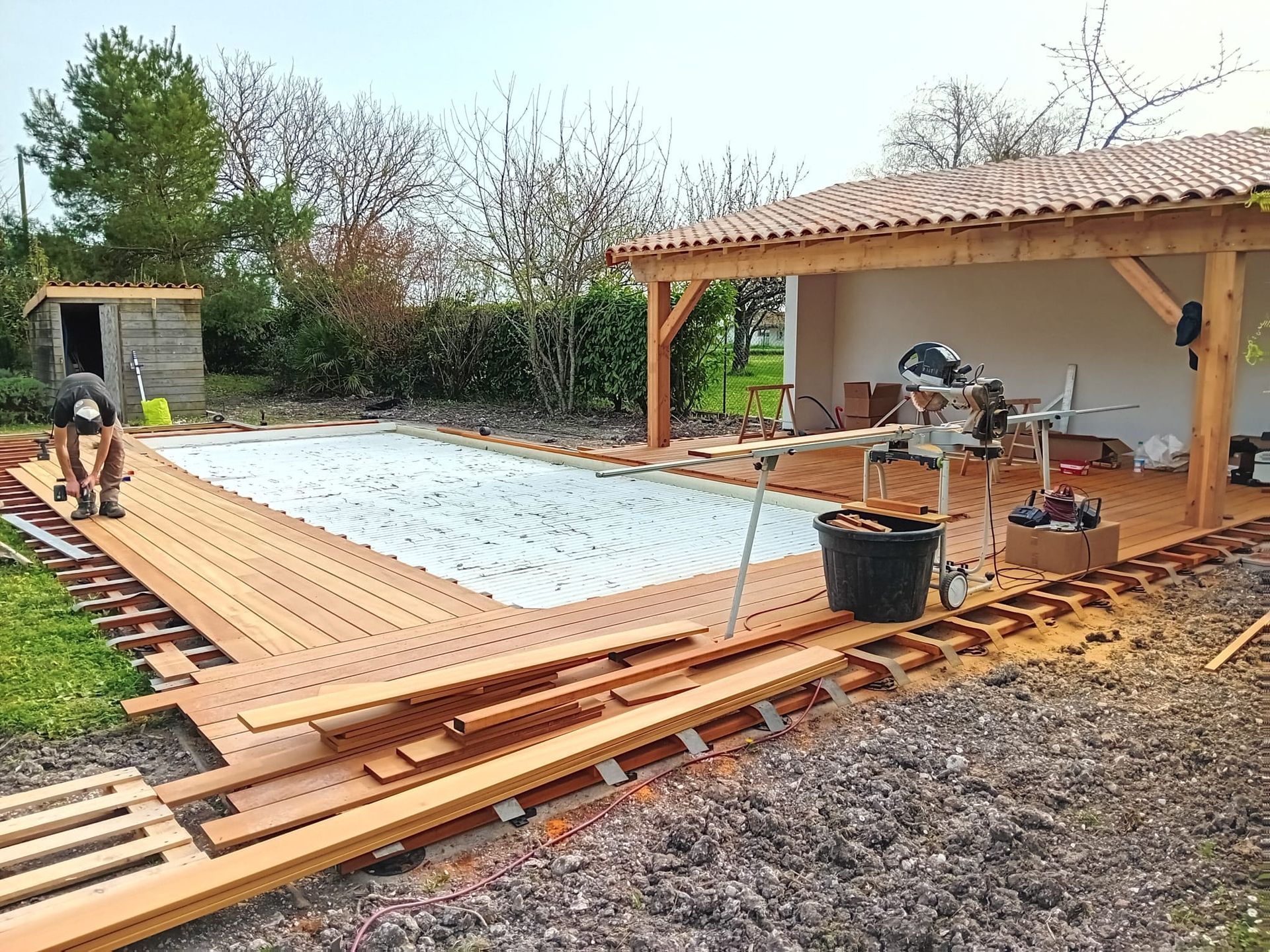 Construction d'une terrasse en bois autour d'une piscine, avec un ouvrier, un abri de jardin et un espace couvert dans une cour.