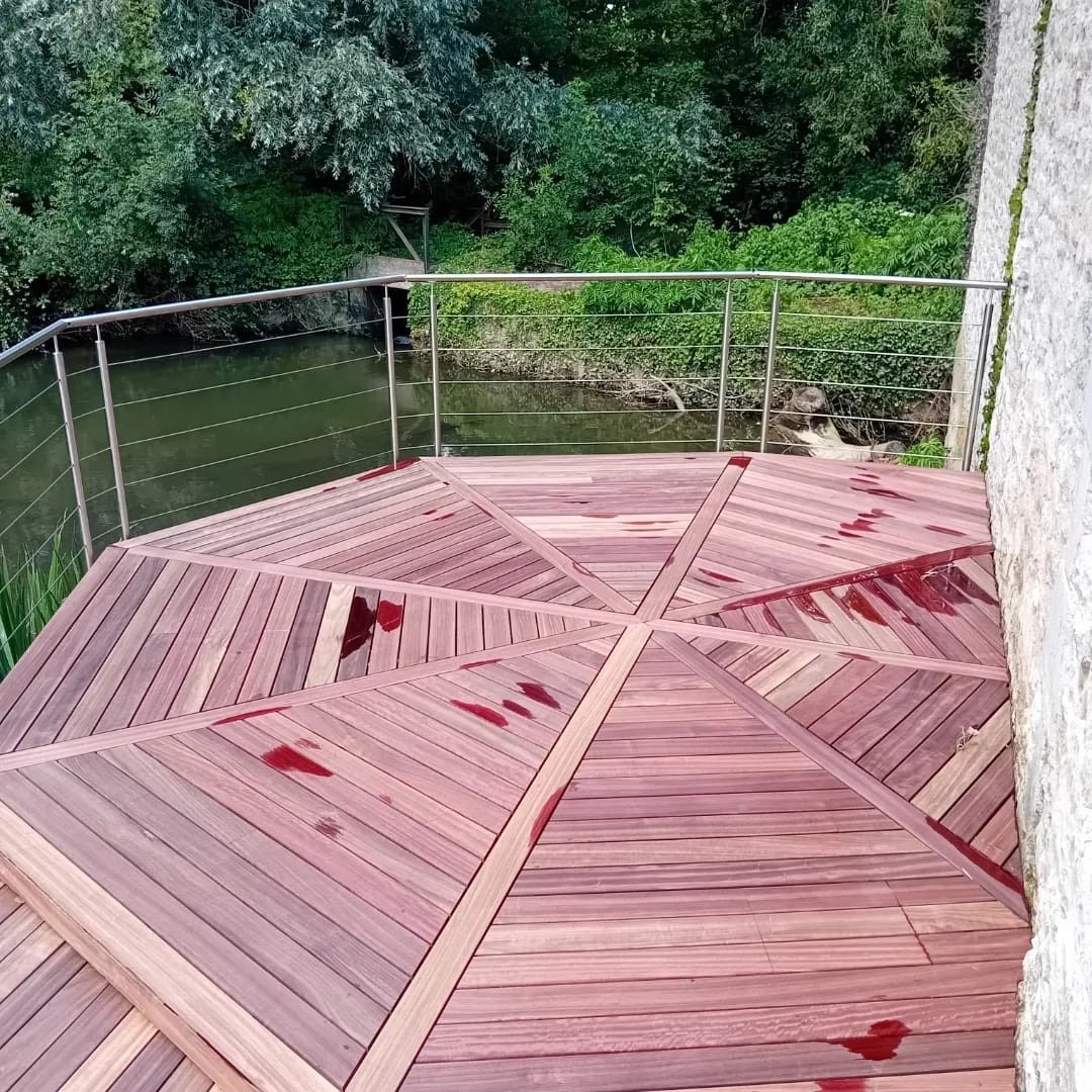Terrasse en bois avec rambarde donnant sur l'eau. Arbres verts en arrière-plan, mur patiné par le temps sur la droite.