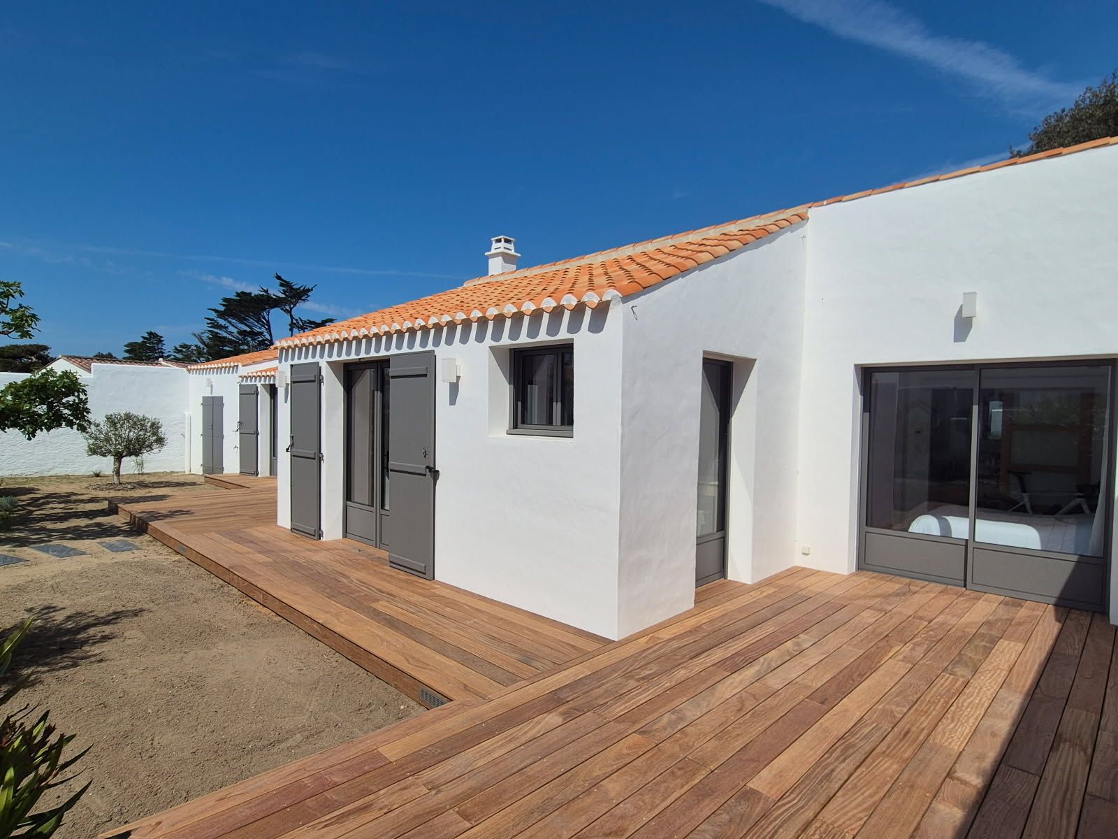 Maison en stuc blanc avec terrasse en bois, volets gris et toit de tuiles en terre cuite.