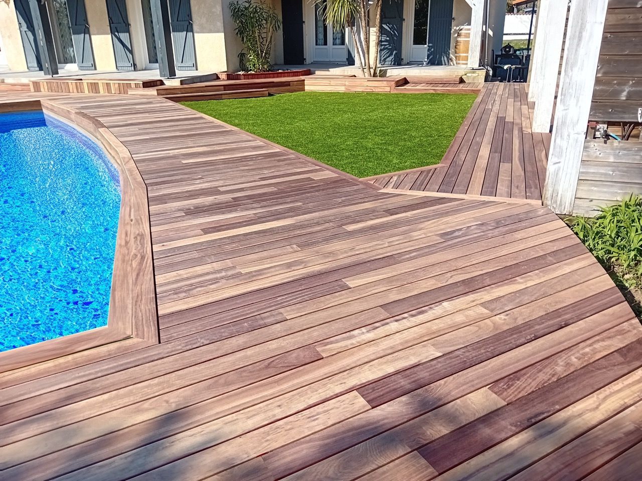La terrasse en bois de la piscine s'incurve autour d'une piscine bleue et mène à une pelouse verte.