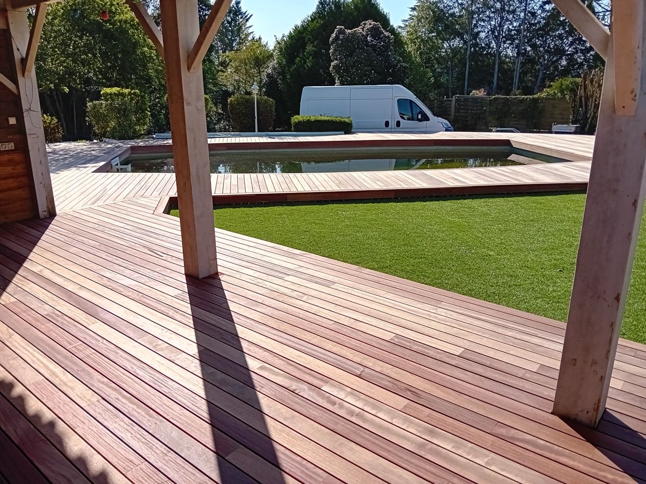Terrasse en bois avec piscine et gazon artificiel ; camionnette blanche en arrière-plan.