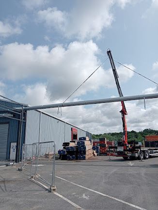 Grue soulevant une longue poutre métallique à côté d’un entrepôt, dans une cour industrielle sous un ciel nuageux