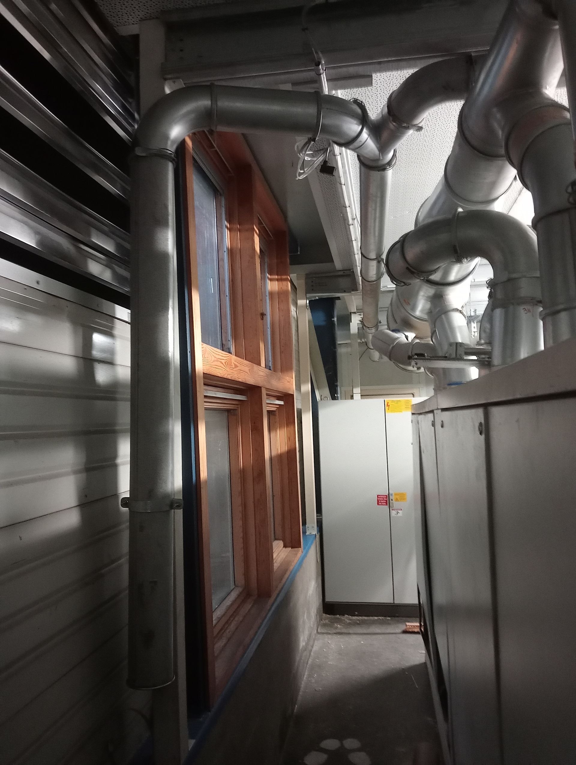 Couloir industriel étroit avec des tuyaux apparents, des conduits métalliques et des armoires électriques le long des murs.