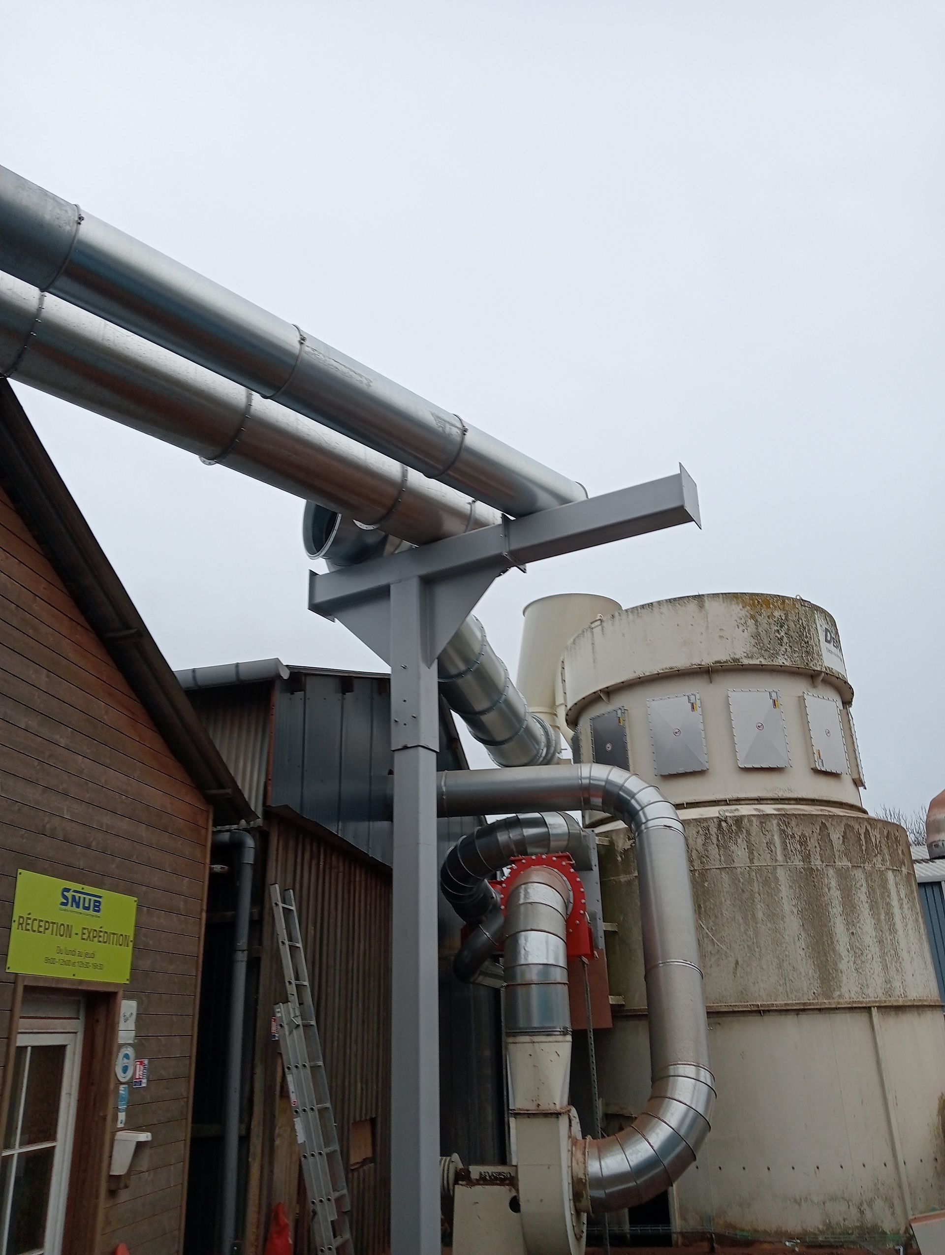 Des tuyaux et des équipements industriels à côté d’un bâtiment en briques et de réservoirs rouillés, à l’extérieur
