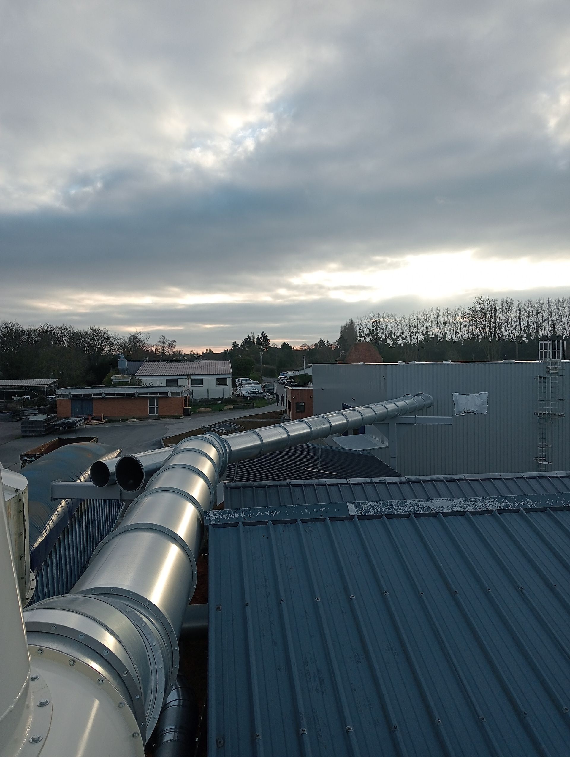 Vue depuis le toit sur des conduits métalliques au-dessus d’un toit bleu en tôle ondulée, sous un ciel nuageux