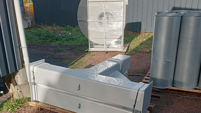 Des panneaux en métal blanc et des parpaings sur une allée en gravier, à côté d’une maison, avec un tonneau gris en arrière-plan