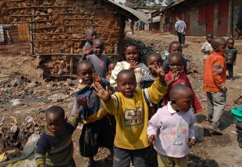 Kinder stehen im Freien auf einem unbefestigten Weg neben rustikalen Gebäuden, lächeln und strecken die Hände Richtung Kamera