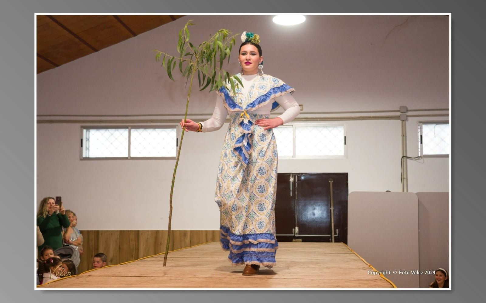 Una mujer con un vestido azul y blanco camina por una pasarela sosteniendo una planta.