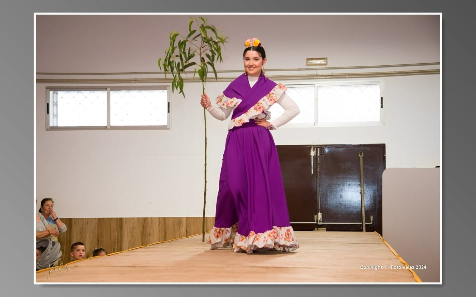 Una mujer con un vestido morado está parada en un escenario sosteniendo una planta.