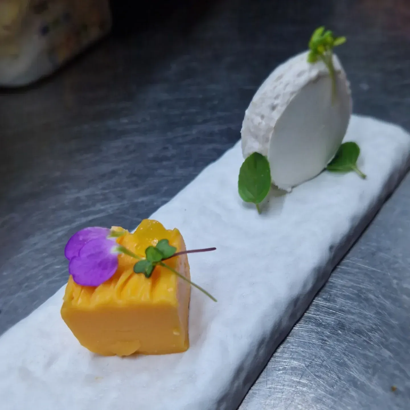 Aperitivo: Cuadrado de naranja y queso blanco sobre un plato rectangular blanco, adornado con flores y hierbas comestibles.