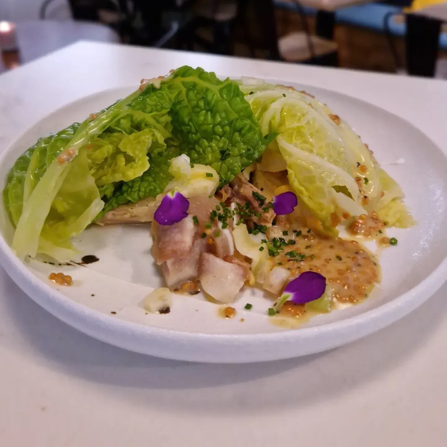 Plato blanco con ensalada compuesta a base de repollo, aderezo, flores comestibles y otros ingredientes.