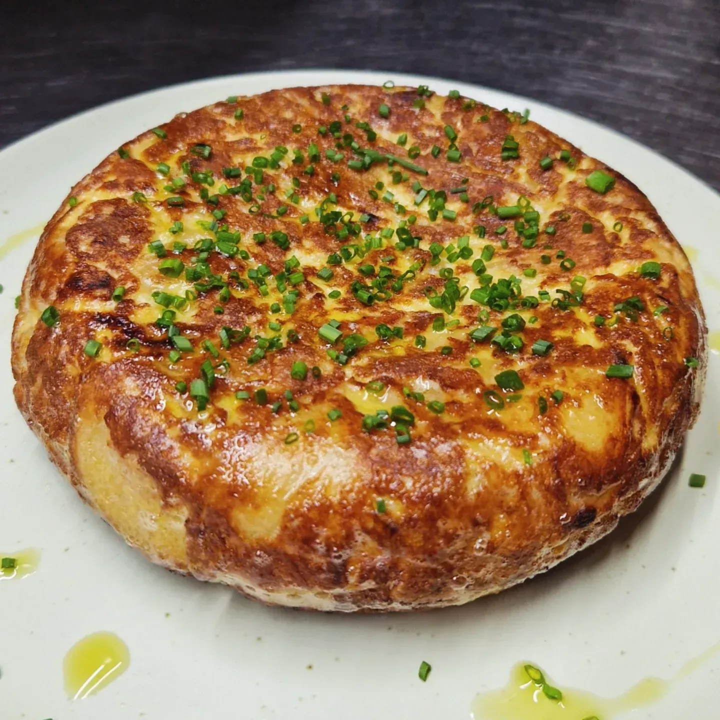 Tortilla española en un plato, dorada con cebollino, rociada con aceite de oliva.