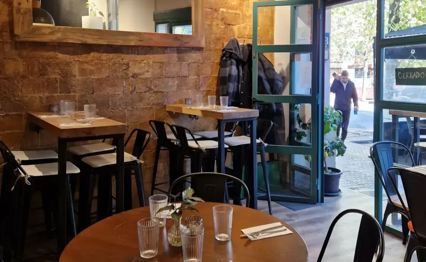 Interior de restaurante con mesas, sillas, pared de ladrillo, puerta abierta y una persona caminando afuera.