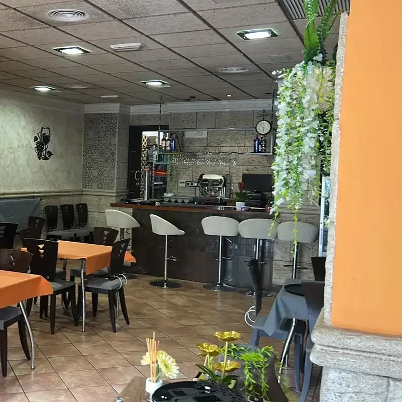 Cafetería vacía con mesas naranjas, sillas negras, una barra y flores blancas colgantes.