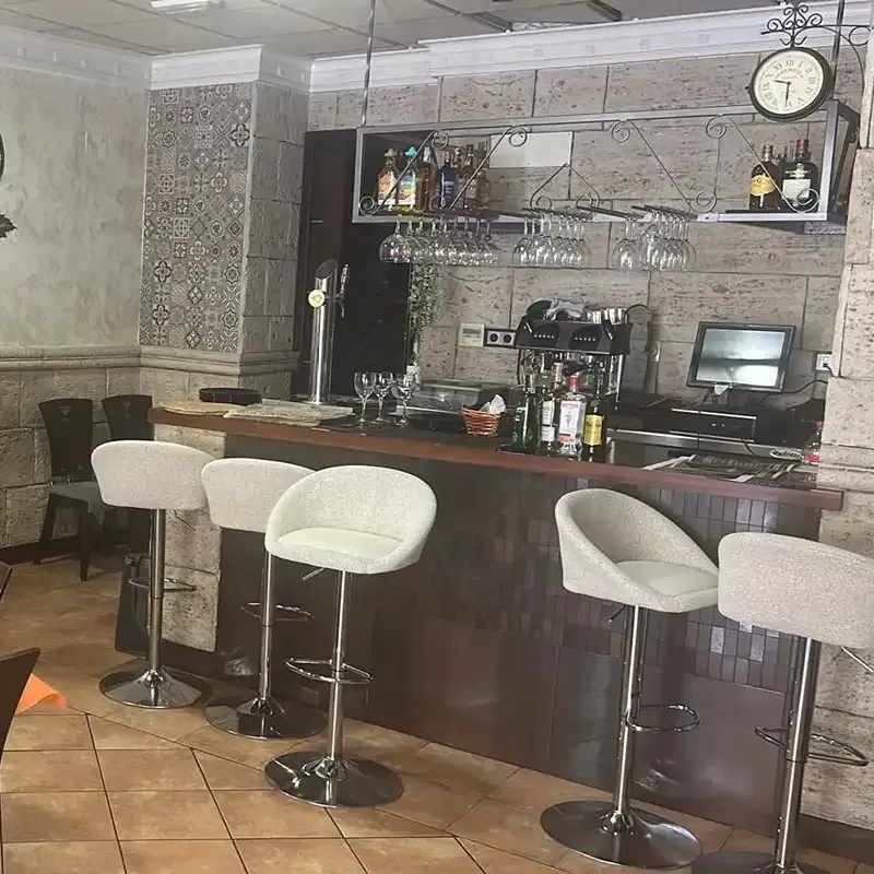 Barra de bar vacía con mostrador de madera oscura, taburetes blancos, copas de vino, botellas y un reloj de pared.