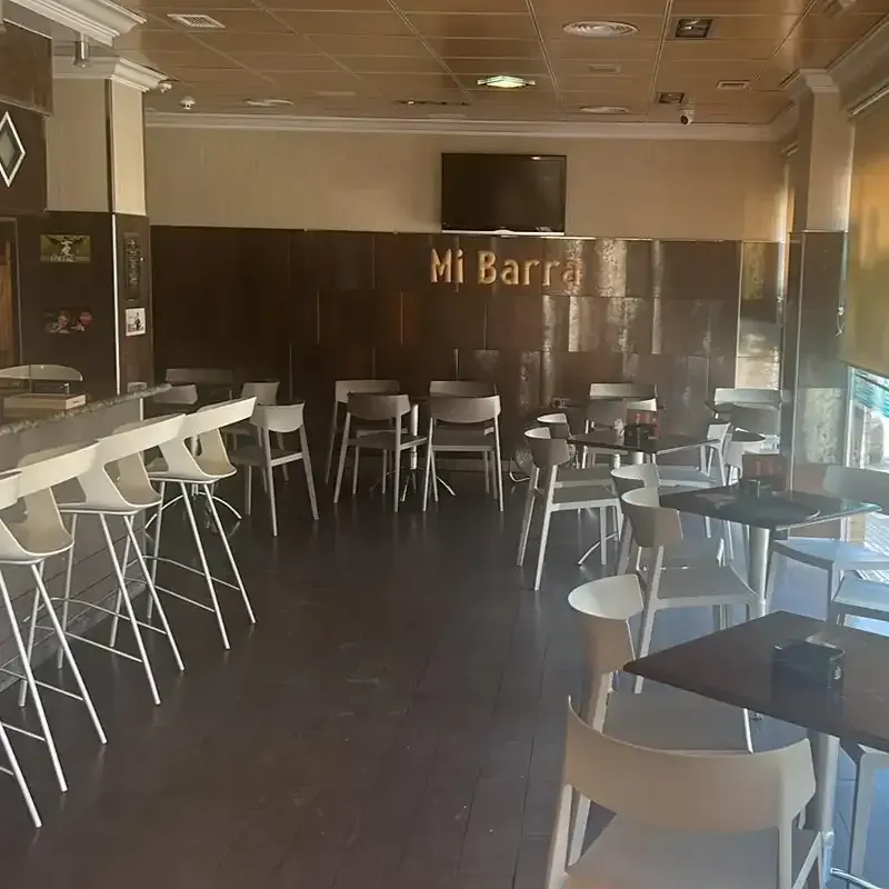 Zona de estar de cafetería vacía con sillas blancas, mesas negras y una barra de madera oscura.