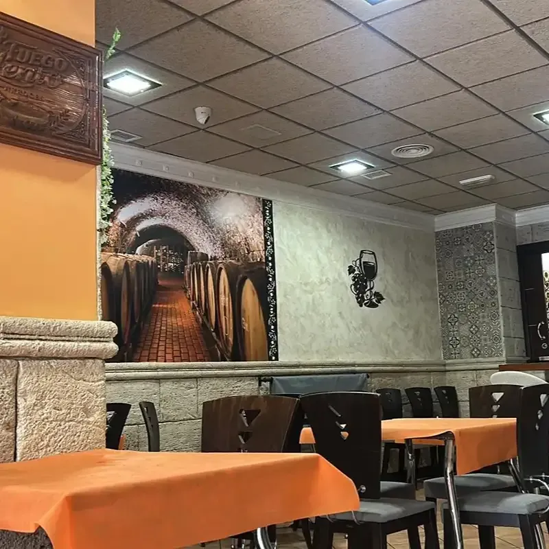 Comedor vacío de un restaurante con manteles naranjas, sillas negras y murales en las paredes bajo un techo de azulejos.