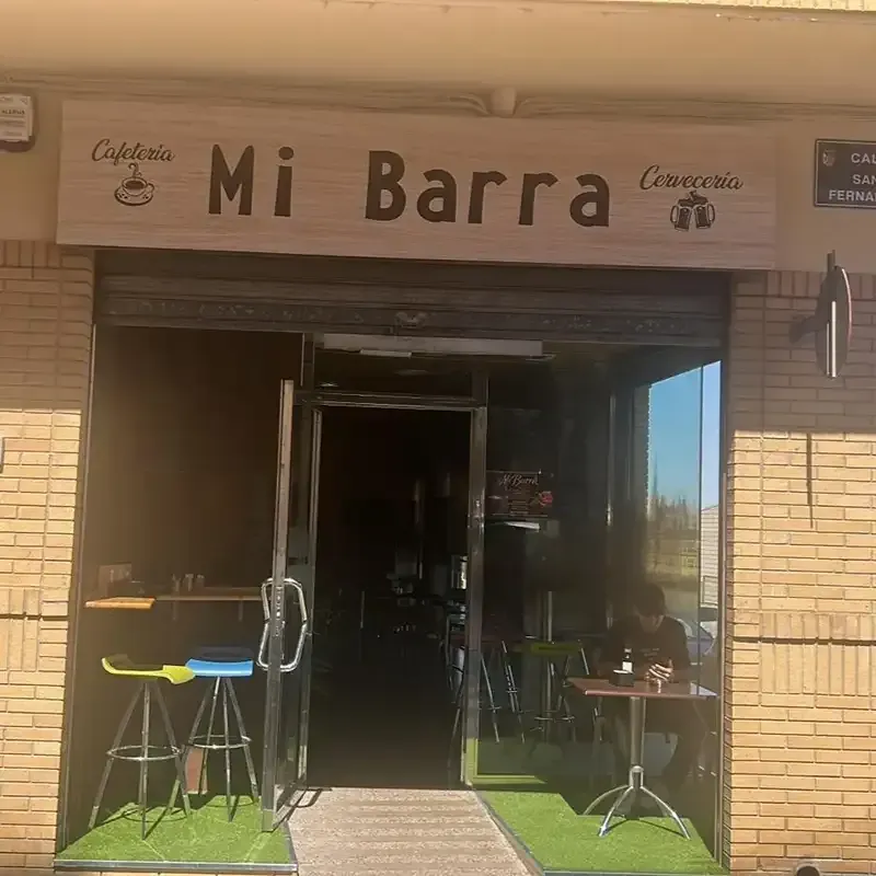 Entrada de la cafetería con letrero de “Mi Barra”, puerta abierta y mesitas en la acera.
