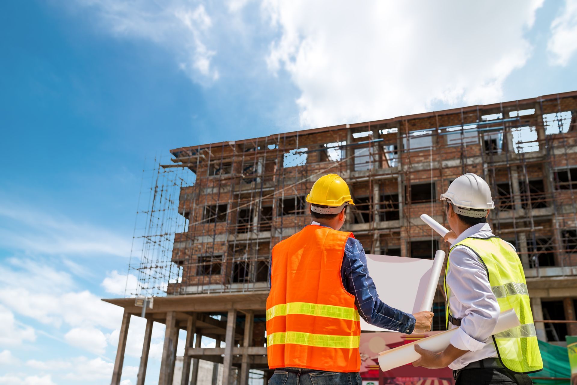 Deux ouvriers du bâtiment, portant des gilets de sécurité et des casques de chantier, examinent des plans sur un chantier.