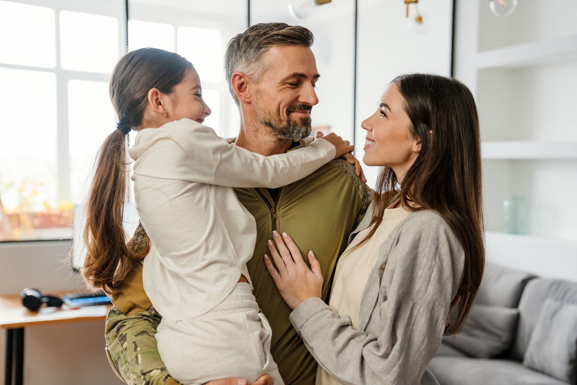 Un homme souriant en chemise verte avec un enfant et une femme dans un salon lumineux.