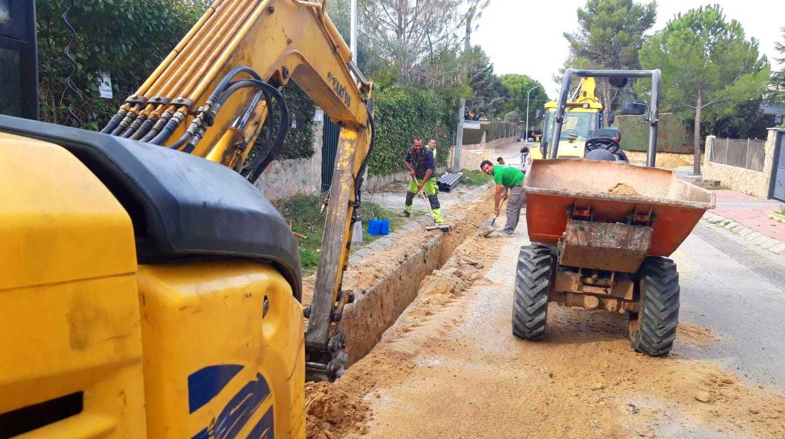 Excavadora amarilla y camión volquete en una calle, hombres trabajando en una zanja.
