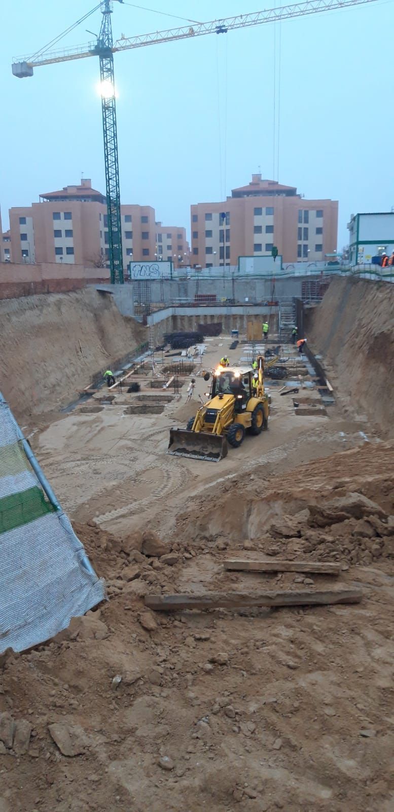 Obra con una excavadora amarilla trabajando en un gran pozo. Una grúa se alza sobre ella, con edificios al fondo.