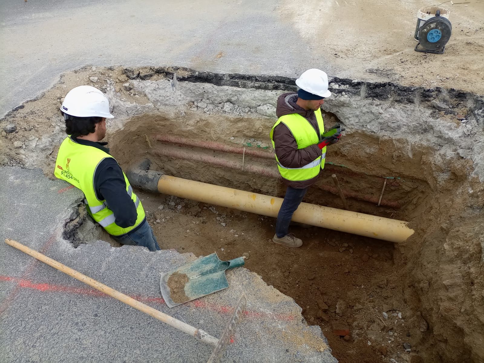 Dos trabajadores con chalecos reflectantes y cascos examinan tuberías en una excavación abierta en una carretera.