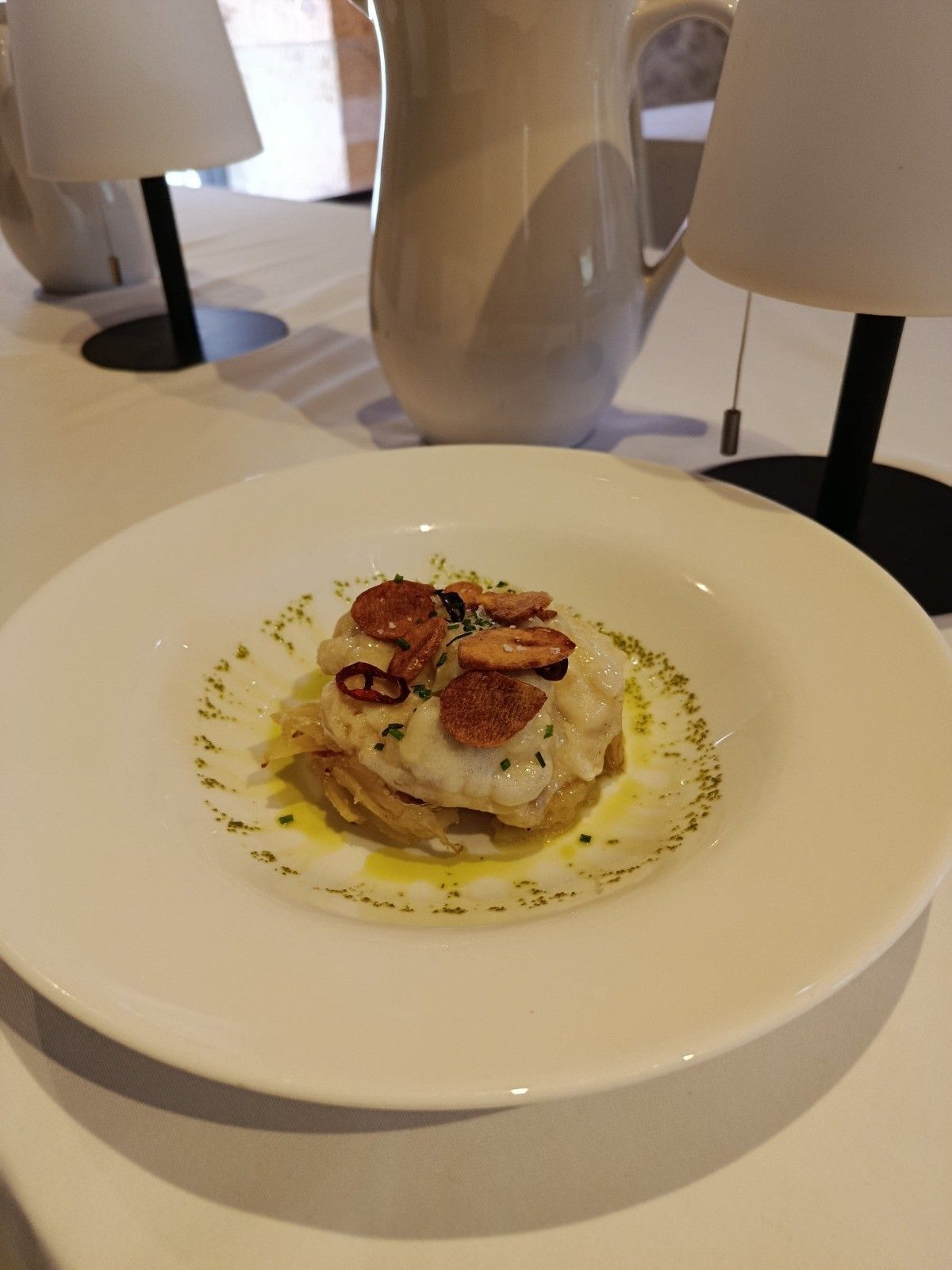 Un plato blanco con pescado cubierto con rodajas de ajo, servido sobre un mantel blanco con dos lámparas y una jarra blanca.