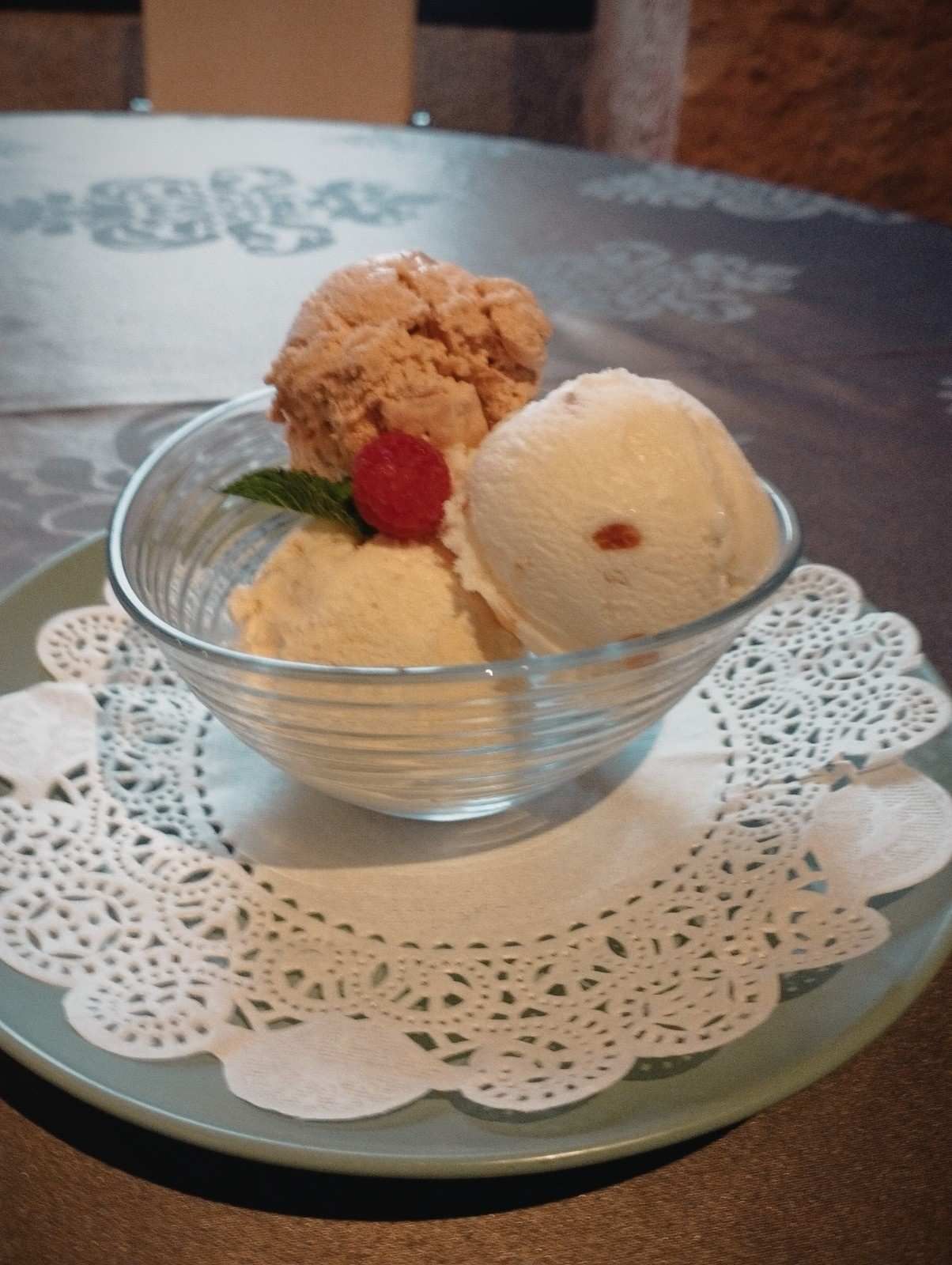 Un bol de cristal con tres bolas de helado sobre un tapete de encaje blanco, adornado con una frambuesa y una hoja de menta.