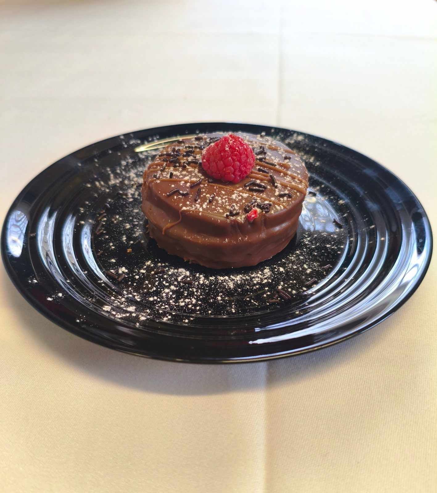 Un pequeño postre redondo, cubierto de chocolate y decorado con una frambuesa fresca, servido en un plato negro acanalado.
