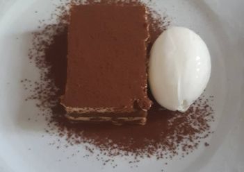 Una porción rectangular de tiramisú cubierta con cacao en polvo, servida en un plato blanco con una bola de helado blanco.