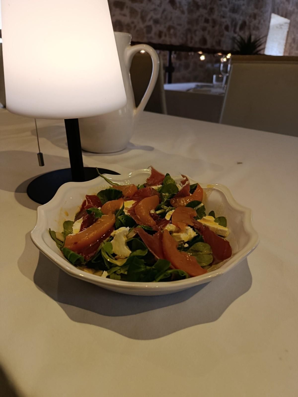 Un pequeño bol de ensalada con verduras, queso y jamón curado, servido sobre un mantel blanco.
