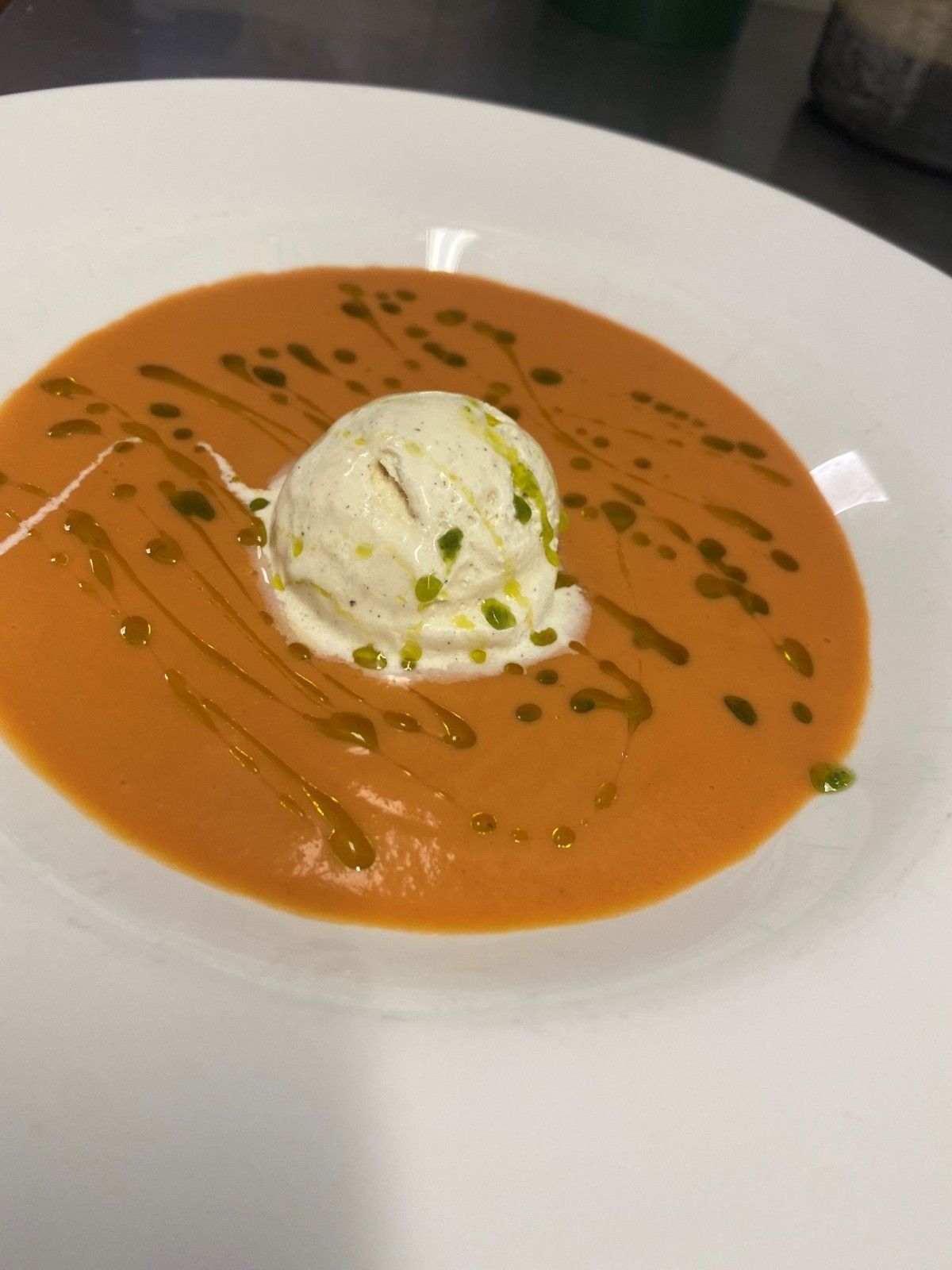 Un tazón de sopa suave de color naranja, coronado con una bola de helado blanco y rociado con aceite de hierbas verdes.