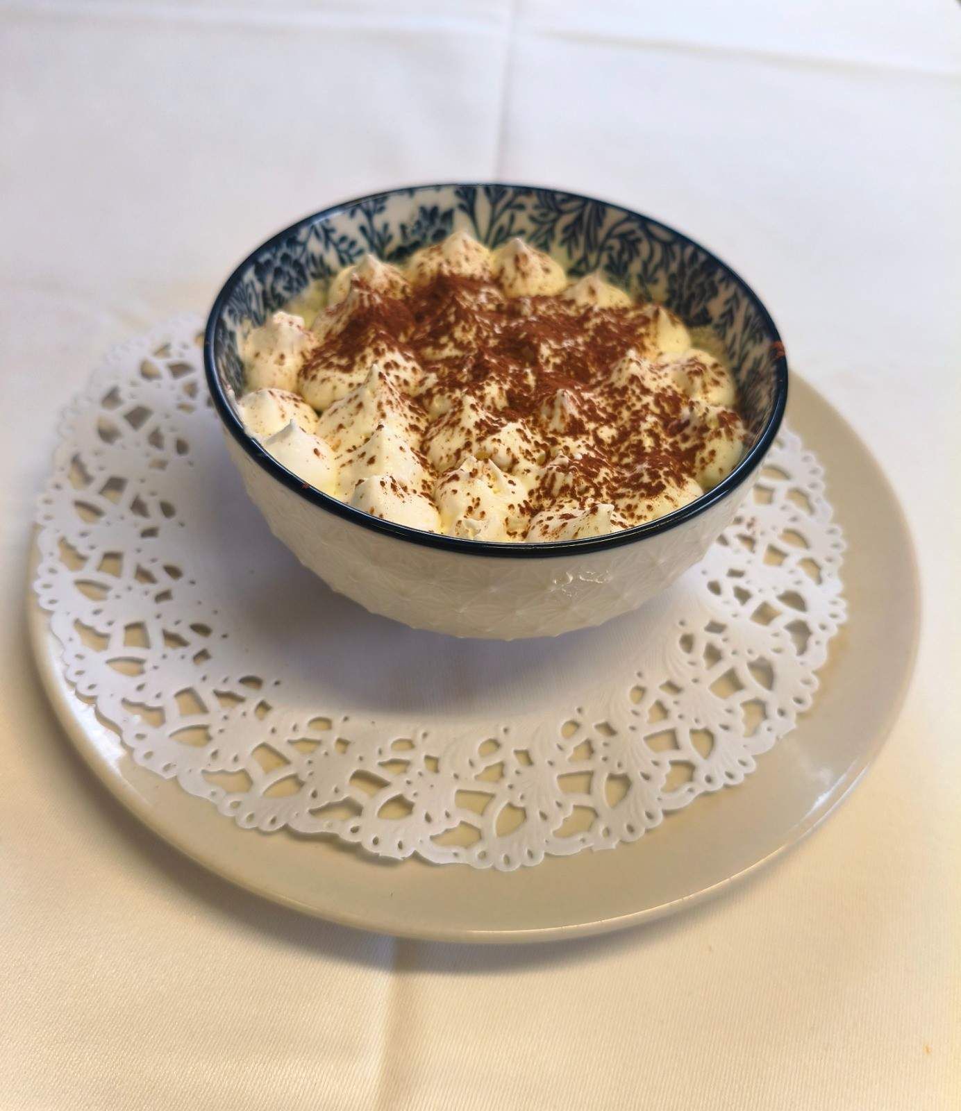 Un bol de tiramisú cremoso espolvoreado con cacao en polvo, servido en un plato blanco con un tapete de papel decorativo.