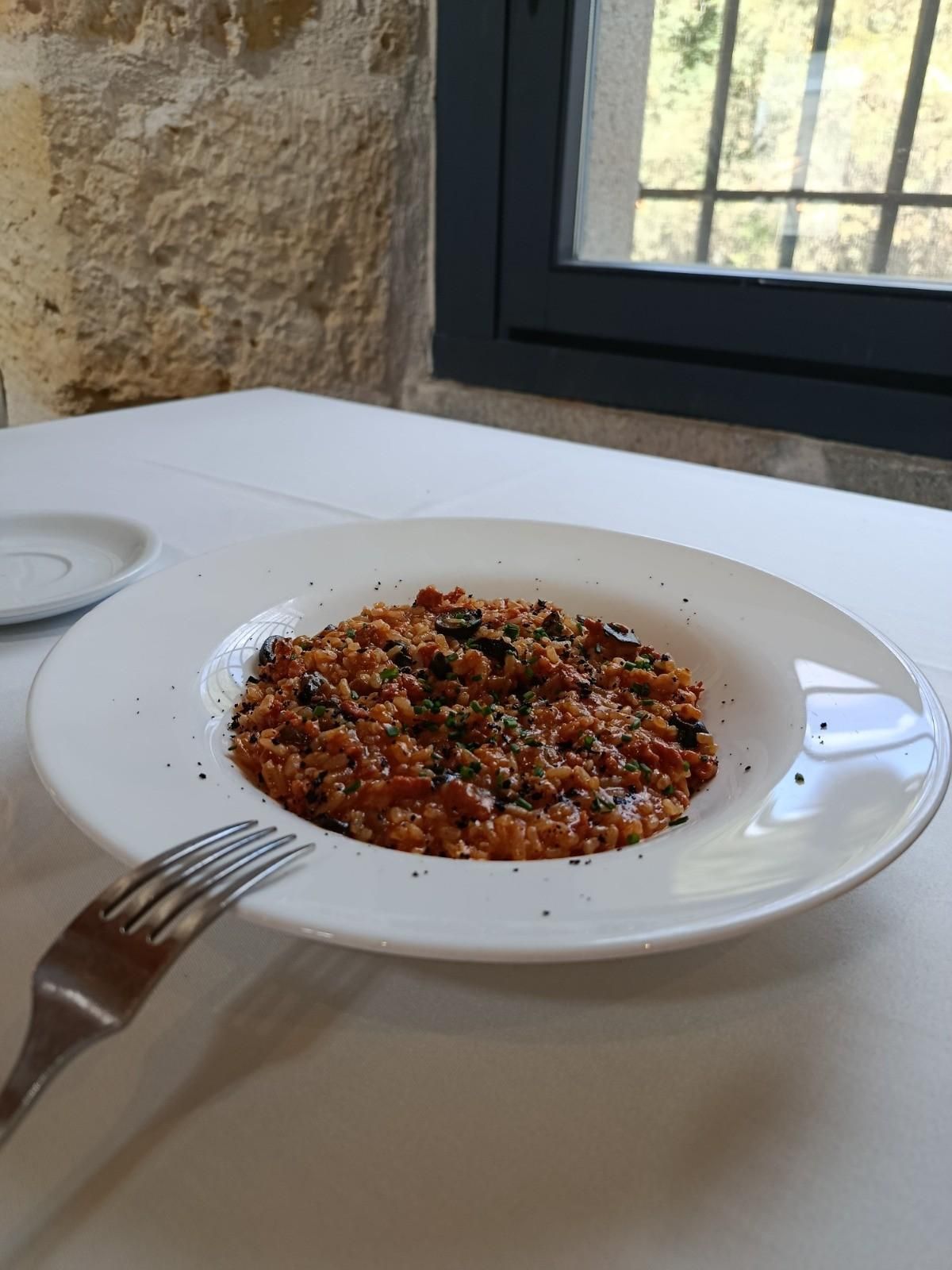 Un bol de risotto a base de tomate con aceitunas negras y hierbas, servido sobre un mantel blanco junto a un tenedor.