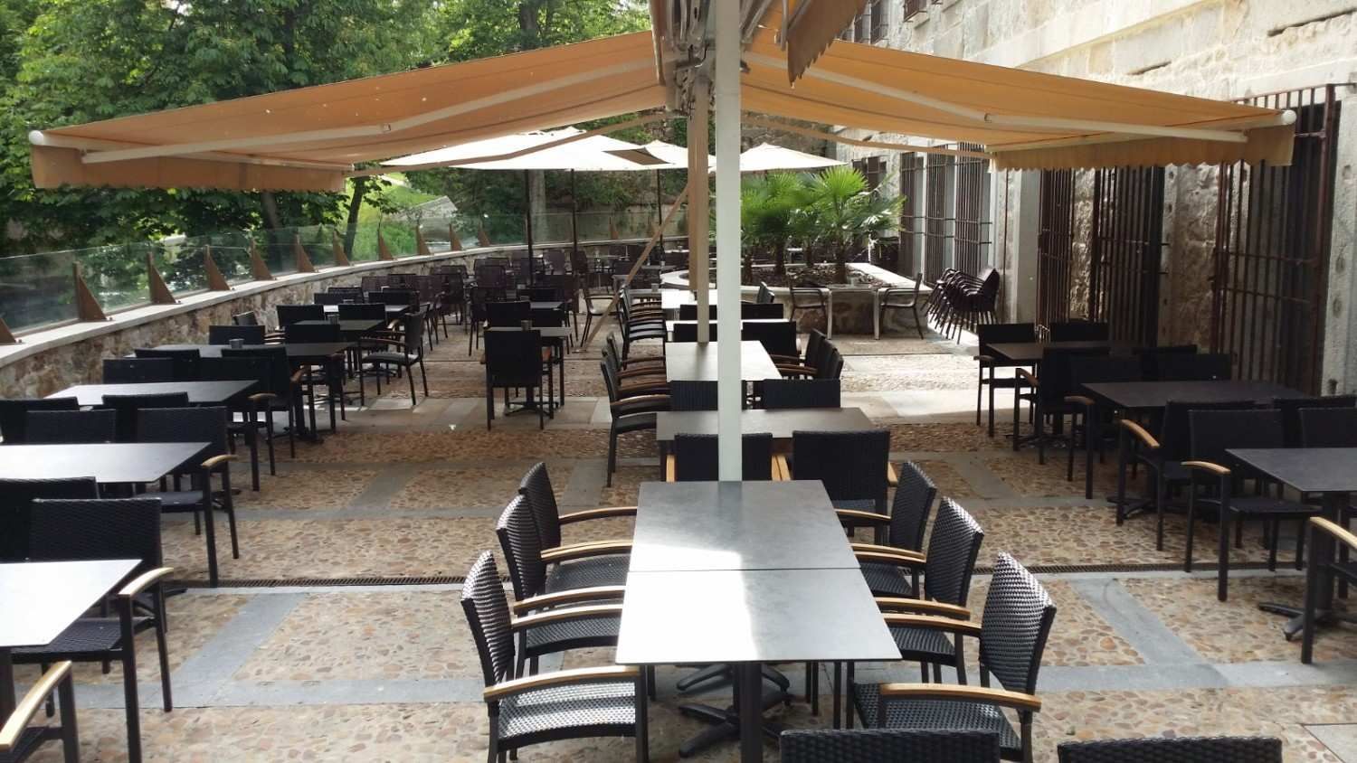 Patio exterior vacío con mesas y sillas oscuras bajo toldos beige en una terraza de piedra pavimentada.