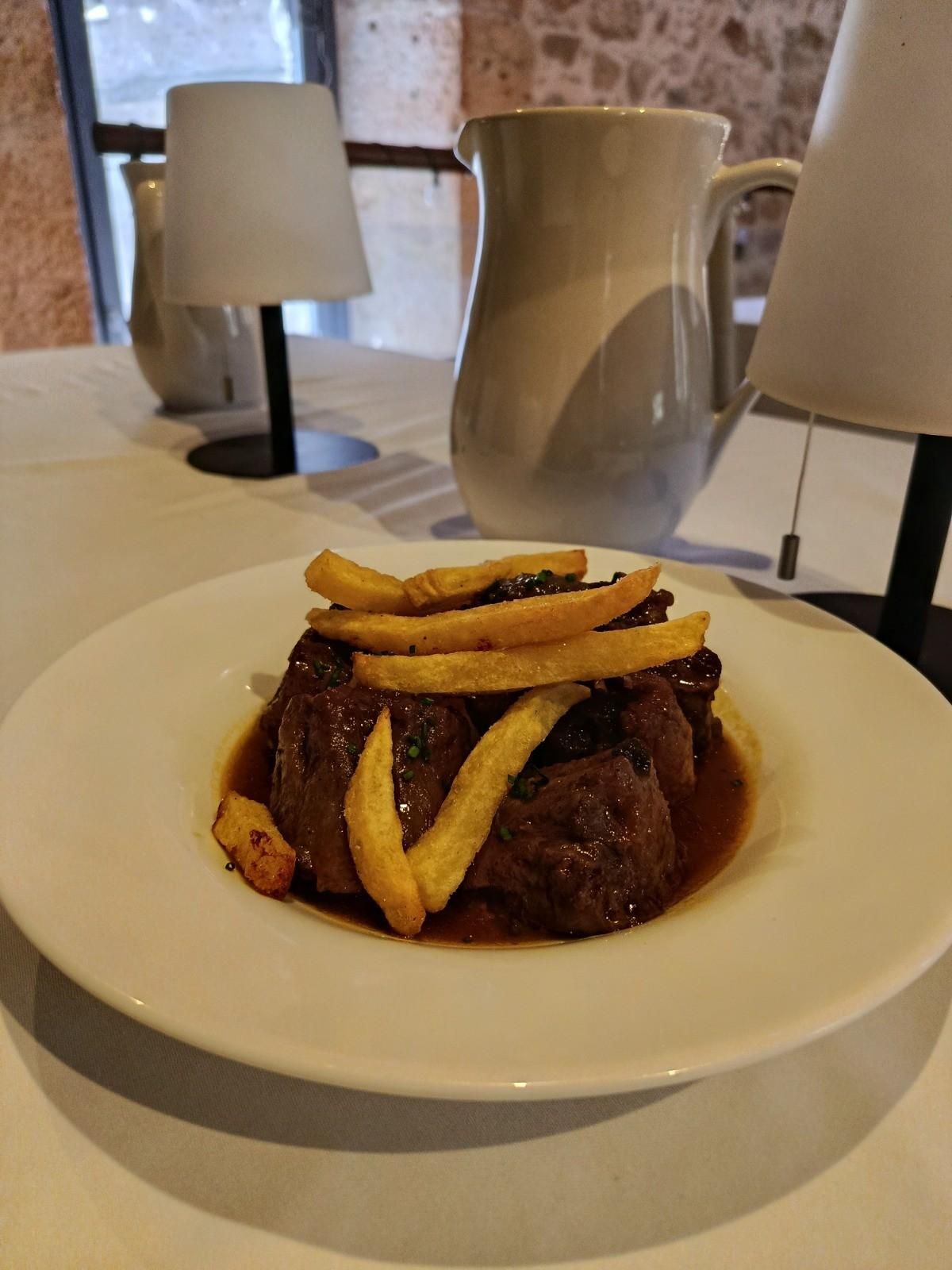 Un plato de carne estofada con patatas fritas, dispuesto sobre una mesa blanca con lámparas y una jarra de agua al fondo.