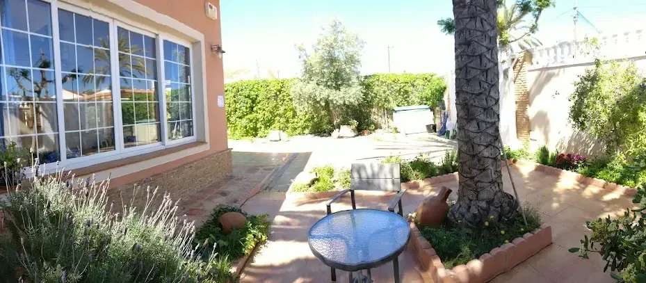 Patio con mesa redonda de cristal, plantas en macetas y tumbonas junto a una casa y un sendero ajardinado.