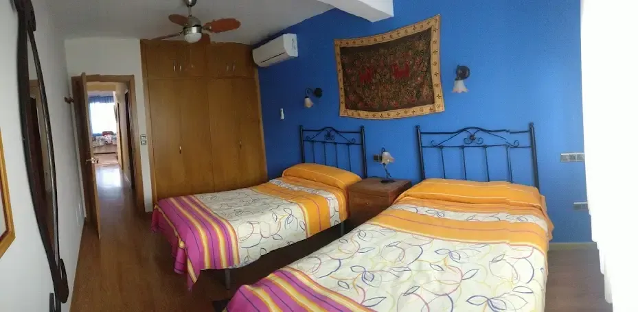 Habitación pequeña con dos camas individuales, paredes azules, armario de madera y una puerta de pasillo al fondo.
