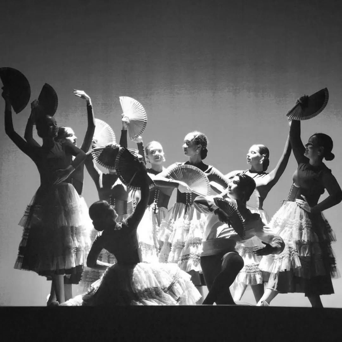 Bailarines con siluetas recortadas y trajes vaporosos posan en un escenario iluminado con luz púrpura, algunos sosteniendo sombreros en alto.