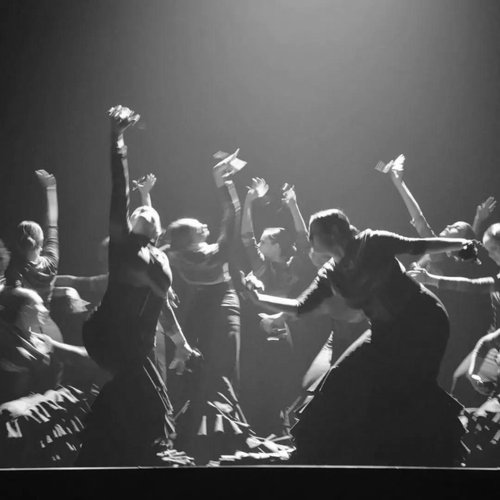 Bailarines vestidos de oscuro se mueven enérgicamente bajo las luces moradas del escenario en una actuación en directo.