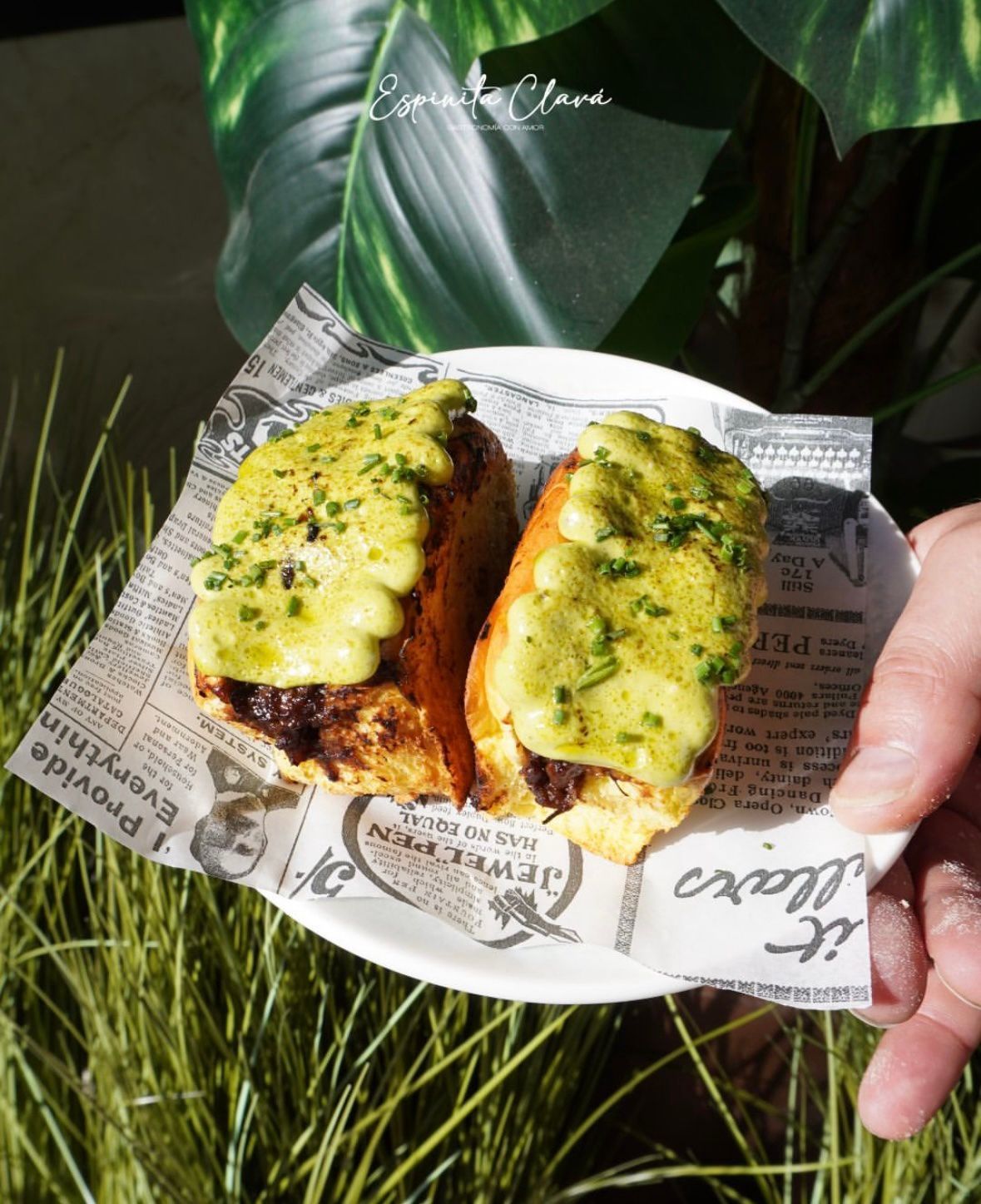 Dos mitades de patata asada con queso sobre papel de periódico en una mano, con hierba y hojas verdes al fondo.