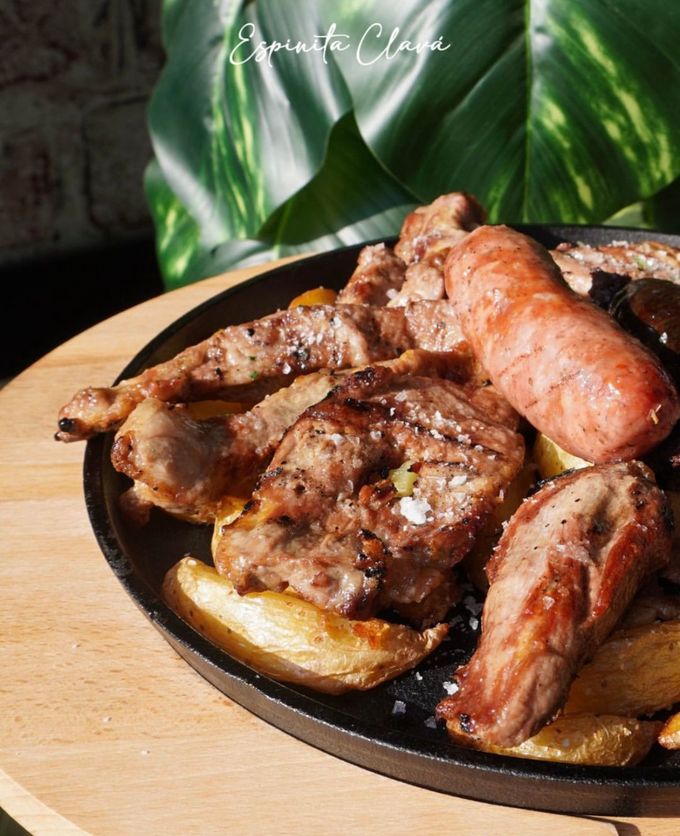 Carnes y patatas a la parrilla en una sartén de hierro fundido sobre una tabla de madera, con hojas verdes al fondo.