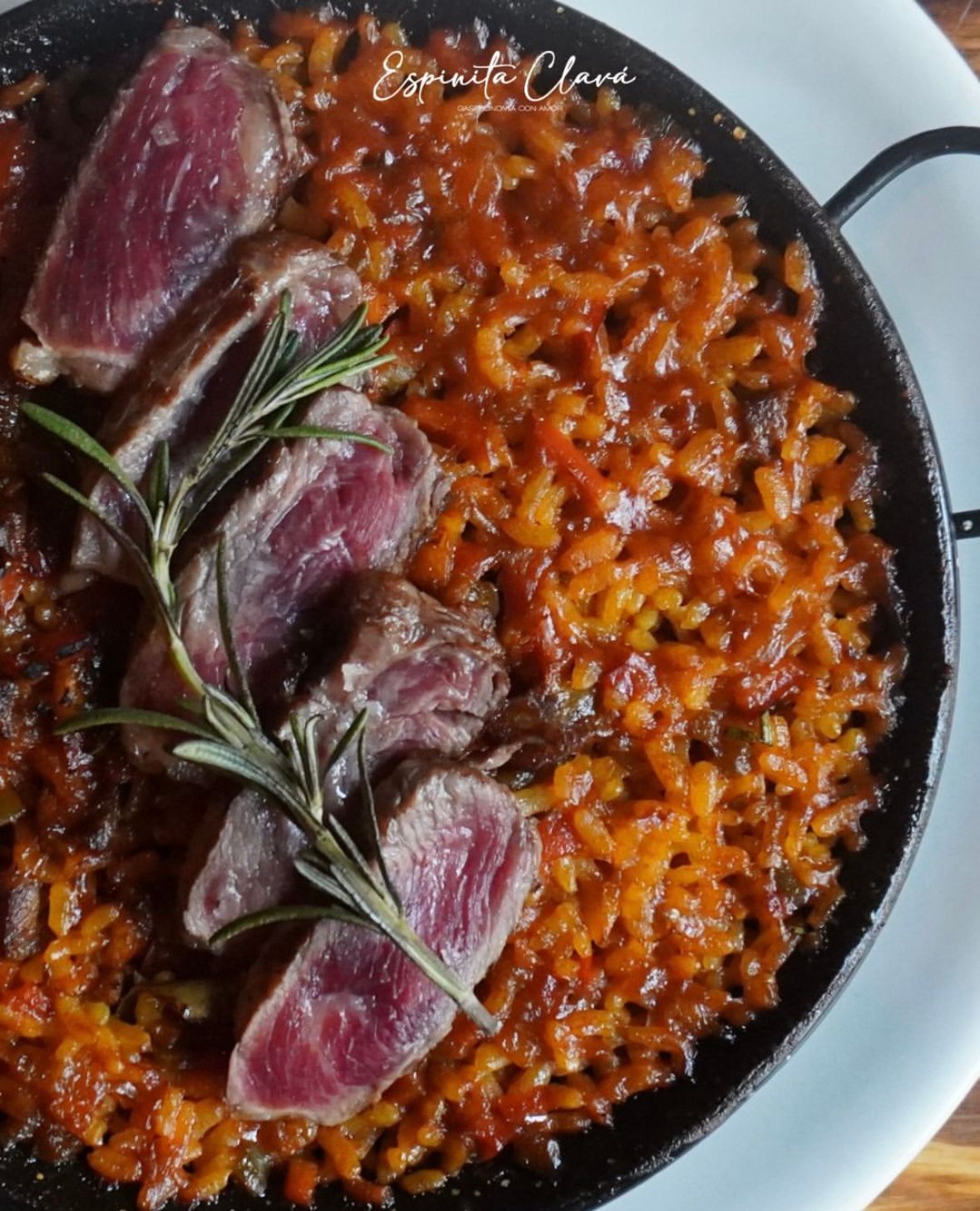 Sartén negra de risotto rojo cubierta con carne en rodajas sellada y romero, servida en un plato blanco.