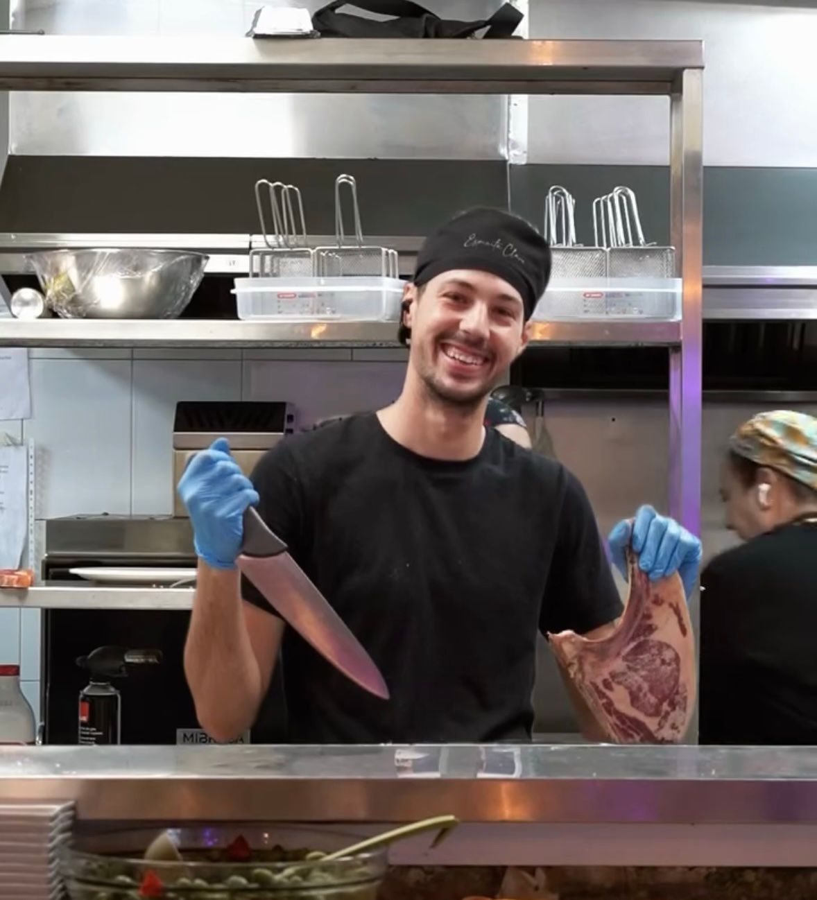 Un chef sonriente, vestido con camisa negra y guantes azules, sostiene un cuchillo y un trozo de carne en una cocina