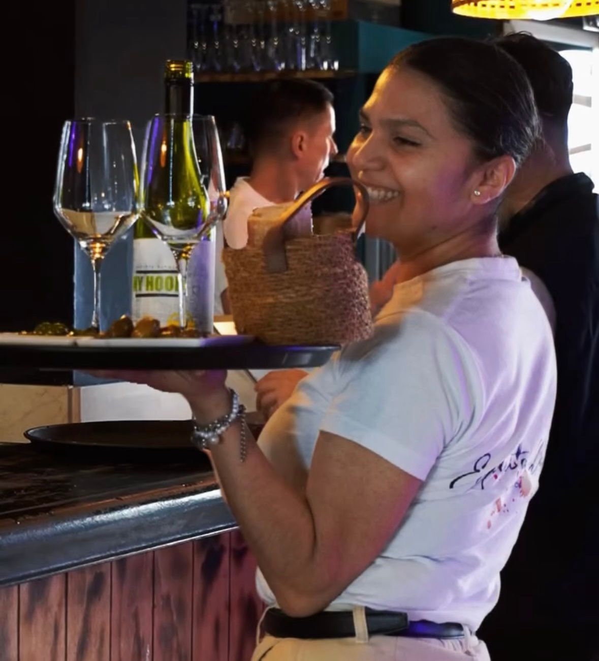 Mujer sonriendo mientras lleva una bandeja con copas de vino y una botella en un bar.