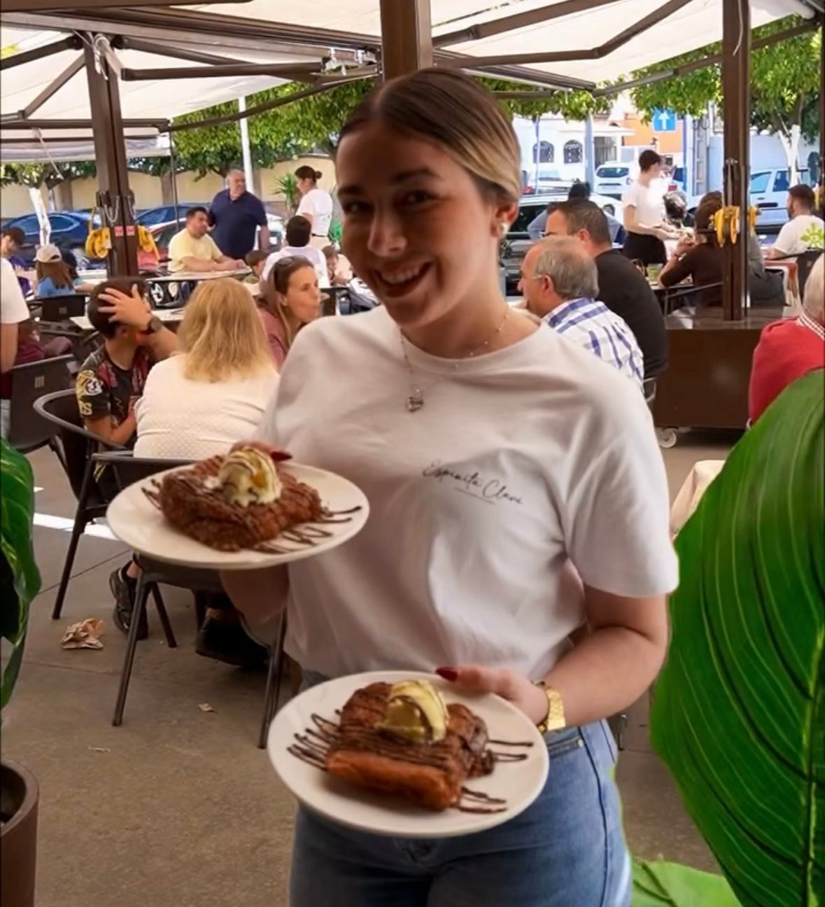 Camarero sonriente con dos platos de postre en una concurrida cafetería al aire libre.