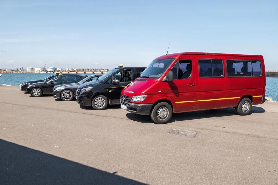 Fourgonnette rouge de passagers garée sur un quai en béton près d'autres voitures, avec une étendue d'eau et un ciel bleu en arrière-plan.