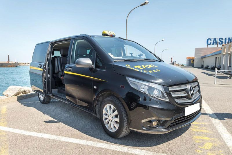 Un taxi Mercedes noir, porte coulissante ouverte, est stationné au bord de l'eau. Un panneau « Taxi » est visible sur le toit, et un casino se trouve en arrière-plan.